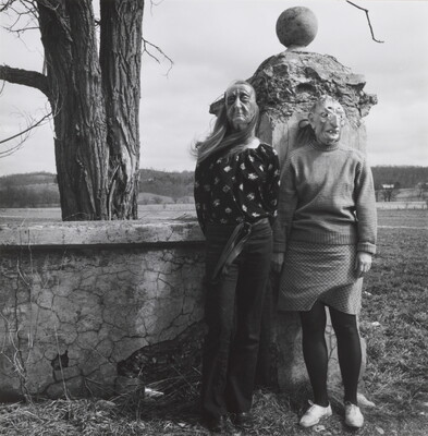 A black-and-white photograph of two women standing in front a stone wall wearing witch-like masks.
