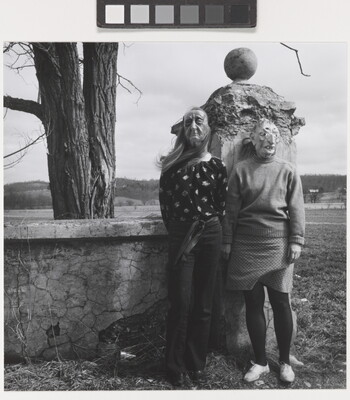 A black-and-white photograph of two women standing in front a stone wall wearing witch-like masks.