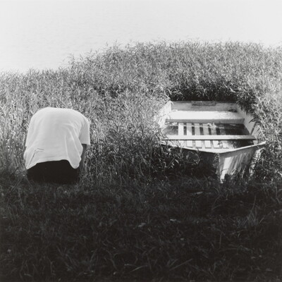 A black-and-white photograph of a hunched over person, back to the camera, next to a rowboat hemmed in by weeds.