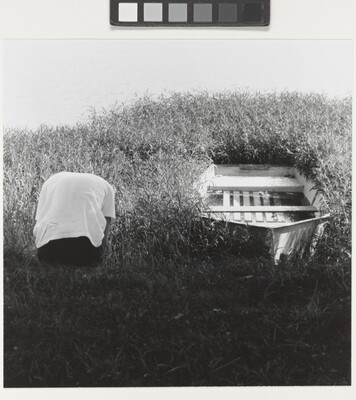 A black-and-white photograph of a hunched over person, back to the camera, next to a rowboat hemmed in by weeds.