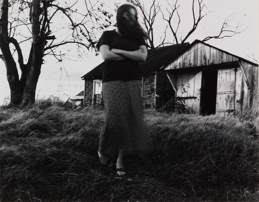 A black-and-white photograph of a White woman, arms crossed and hair blowing in her face, walking toward the viewer from an old wood and brick structure.