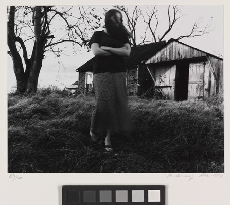 A black-and-white photograph of a White woman, arms crossed and hair blowing in her face, walking toward the viewer from an old wood and brick structure.