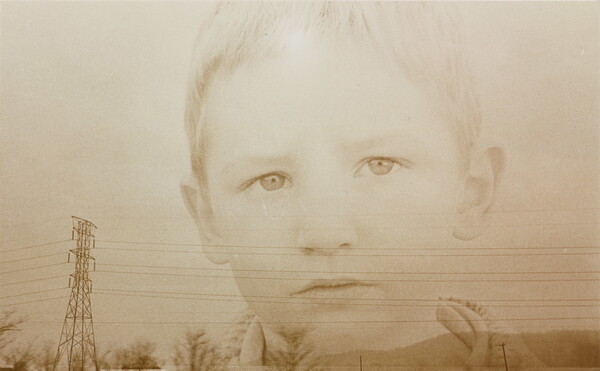 A sepia-toned double-exposed photograph of the face of a young White child over a landscape of powerlines and hills.