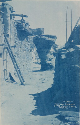 A blue-tinted photograph of a walkway between multi-story pueblo buildings.
