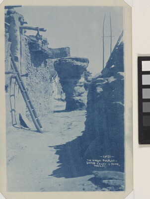 A blue-tinted photograph of a walkway between multi-story pueblo buildings.