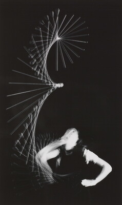 A black-and white photograph of a White person looking up at a baton that is twirling through the air; the baton and the person's arm are blurred indicating motion.