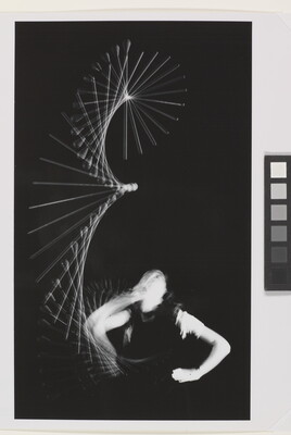 A black-and white photograph of a White person looking up at a baton that is twirling through the air; the baton and the person's arm are blurred indicating motion.