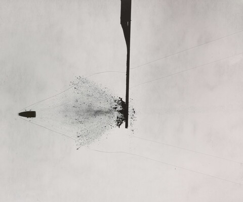 A black-and white photograph of a bullet that has busted through a solid object in the center; a splatter pattern coming from the left of the object follows the bullet.