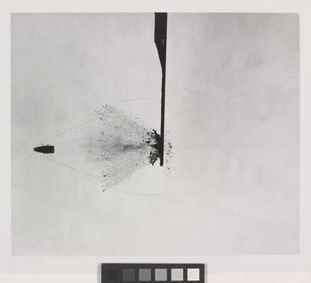 A black-and white photograph of a bullet that has busted through a solid object in the center; a splatter pattern coming from the left of the object follows the bullet.