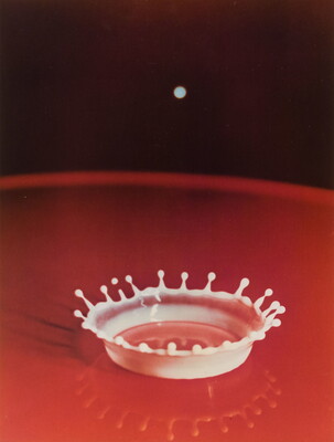A color photograph of a red liquid; a splash of white that resembles a crown rises up and reflects in the liquid.