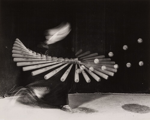 A black-and-white photograph of a blurred person swinging a baseball bat and hitting a ball; the arc of multiple bats and balls indicate motion.