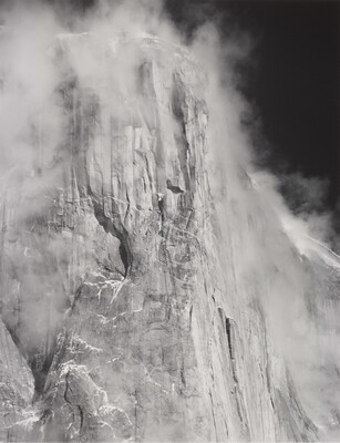 A black-and-white photograph of the side of a tall cliff face surrounded by clouds.