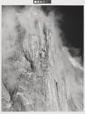 A black-and-white photograph of the side of a tall cliff face surrounded by clouds.