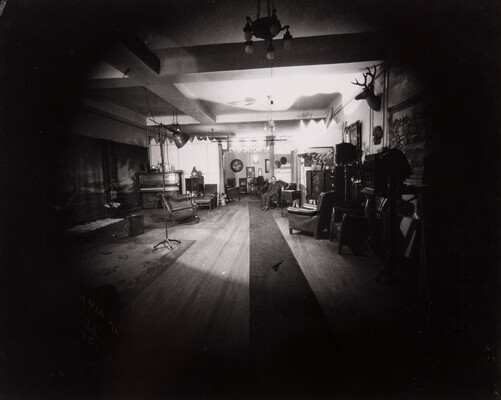 A dark black-and-white photograph of a living room with a spotlight on a man seated in an armchair in a far corner.