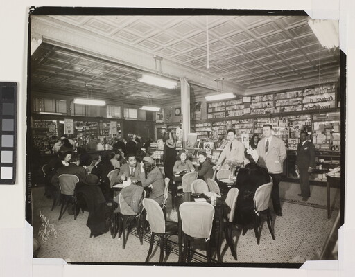 A black-and-white photograph of the interior of a soda fountain filled with a crowd of men and woman socializing.