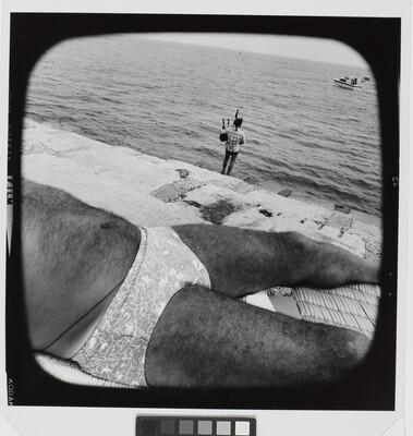 A black-and-white photograph of the hairy torso of a man wearing a brief-style swimsuit sunbathing near a cement ledge by water where a fully-clothed person plays bagpipes in the background.