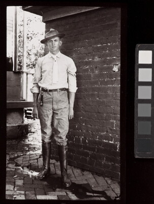 A black-and-white photograph of a young White man in a brimmed hat, light shirt and tie, pants, and tall boots standing in front of brick wall.