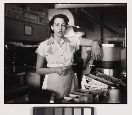 A black-and-white photograph of a White woman in a waitress uniform standing behind a diner counter with an arm propped on drink dispenser.