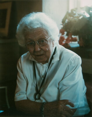 A color photograph of an older White woman with white hair, glasses, and a white blouse leaning forward with her arms crossed.