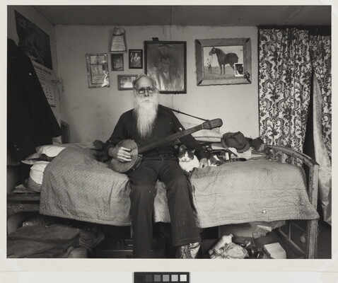 A black-and-white photograph of an elderly White man with a long white beard sitting on a bed in a cozy bedroom holding a banjo with one hand, the other hand around a cat lying next to him.