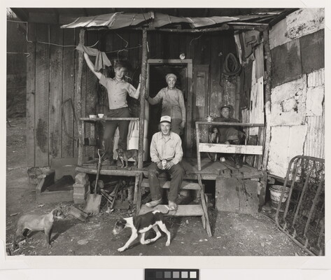 A black-and-white photograph of two people standing, two sitting, and a small dog on the porch of a shack as two dogs play in the dirt yard in front of them.