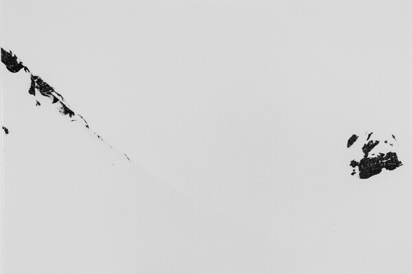 A black-and-white photograph of mountains totally covered with snow except for dark rocky formations in the upper left and center right.