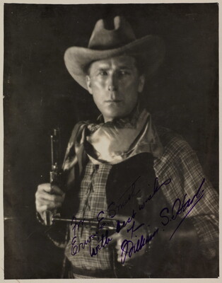 A black-and-white photograph of a White man in western attire holding a pistol in each hand, an autograph in blue in across the bottom.