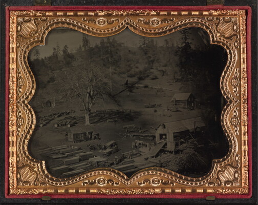 A black-and-white photograph in a decorative gold frame of several buildings and stacks of sheets of wood in a dirt yard.