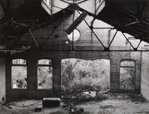 A black-and-white photograph of the inside of an abandoned concrete building with missing windows and debris scattered on the floor.