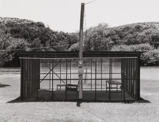 A black-and-white photograph of a screened shelter, a table and chair inside, next to a calm river.