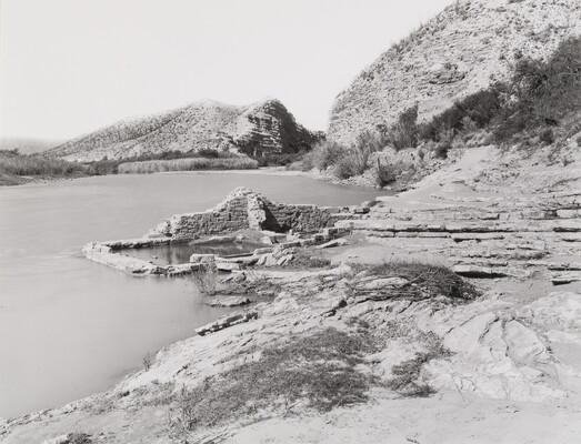 A black-and-white photograph of a body of water surrounded by rocky hillsides and mountains; a crumbled stone building sits on the shore.