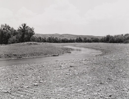 A black-and-white photograph of a river lined with rocks and gravel with many trees in the distance.
