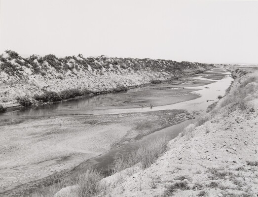 A black-and-white photograph of a near dry river bed with just streams of water flowing between the banks.