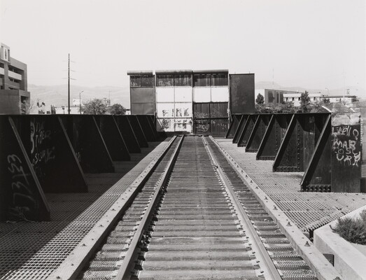A black-and-white photograph of a train track, metal barriers on both sides, that ends at a tall metal gate covered in graffiti.