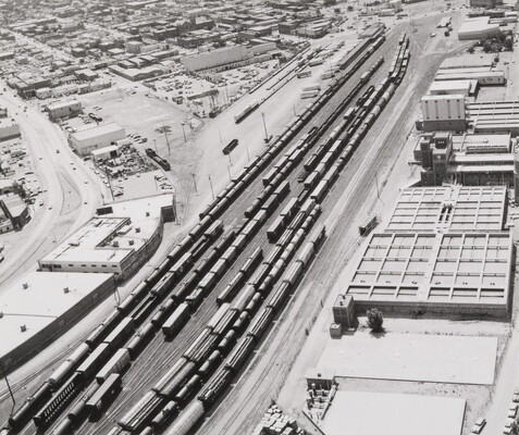 A black-and-white aerial photograph of a railroad yard full of parked trains; warehouses and buildings surround the yard on both sides.