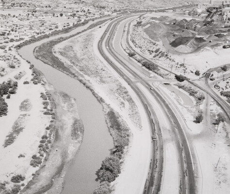 A black-and-white aerial photograph of a road along one side of a river; shrubs and trees dot the sandy landscape.