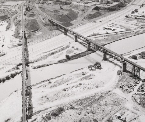 A black-and-white aerial photograph of two railroad bridges spanning across a river and its banks.