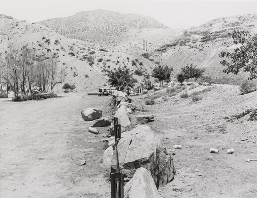 A black-and-white photograph of a dirt road next to a line of boulders and fencing that bisect the scrubby hills.