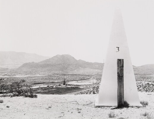 A black-and-white photograph of a tall, white obelisk in a desert, the number three written on one side, mountains and railroad tracks in the distance.