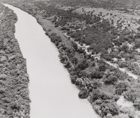 A black-and-white aerial photograph of a river, both shores dotted with trees and shrubs.
