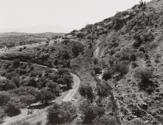 A black-and-white photograph of of a fence bisecting hills covered with scrubby vegetation; a curving dirt road winds around the hills on one side.