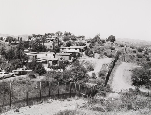 A black-and-white photograph of a fence undulating over hills; a dirt road on one side, low houses and cars on the other.