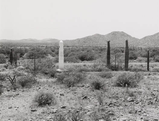 A black-and-white photograph of a white obelisk in a rocky desert near tall cacti and mountains in the distance.