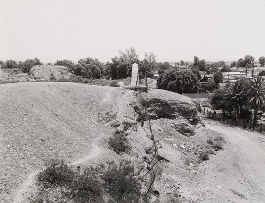 A black-and-white photograph of a white obelisk on a sandy eroding hillside with palm trees and buildings in the distance.
