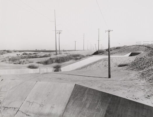 A black-and-white photograph of a concrete channel winding through sandy hills toward a tunnel; telephone poles and scrubby vegetation dot the hills.