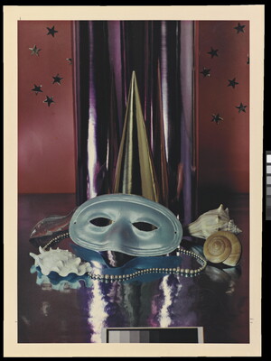 A color photograph of a simple party mask, Mardi Gras beads, and seashells in front of a reflective silver cylinder.
