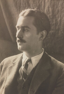 A sepia-toned photograph of a White man with a moustache wearing a dark suit, white shirt, and striped tie looking to the side.