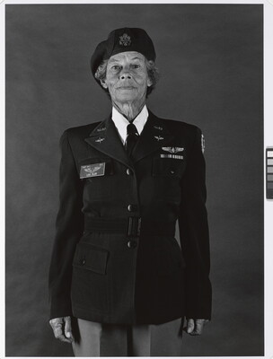 A black-and-white photograph of an older White woman with a wrinkled face wearing a beret and military uniform, standing straight and looking at the viewer.