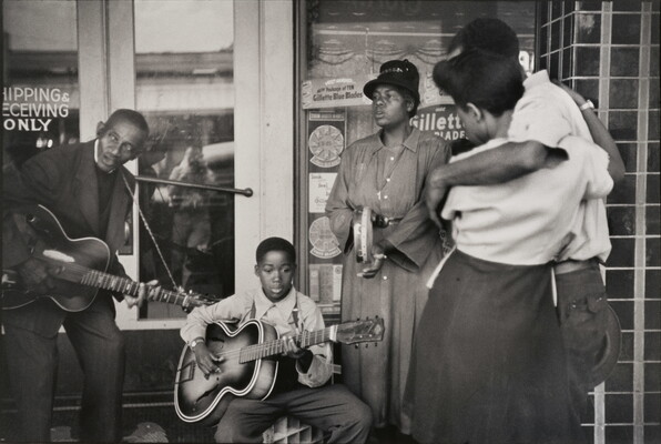 A black-and-white photograph of Black adults and boy playing instruments in front of a storefront while a Black couple watches.