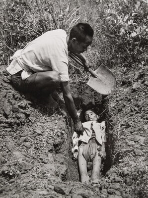 A black-and-white photograph of an Asian man holding a shovel crouched down next to a freshly dug grave containing a emaciated body.